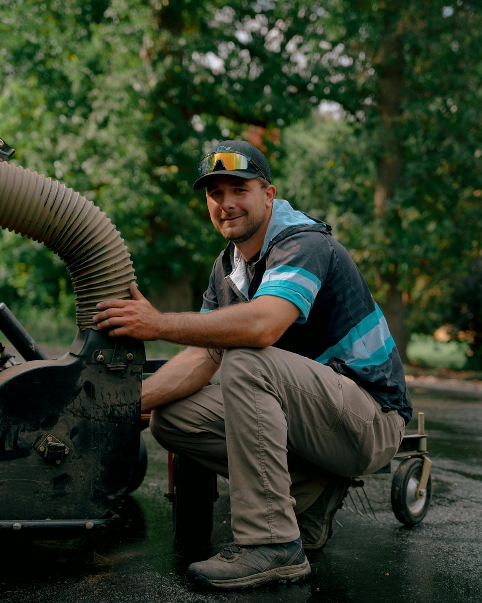 Landscaper crouched beside a leaf vacuum on a tree-lined street